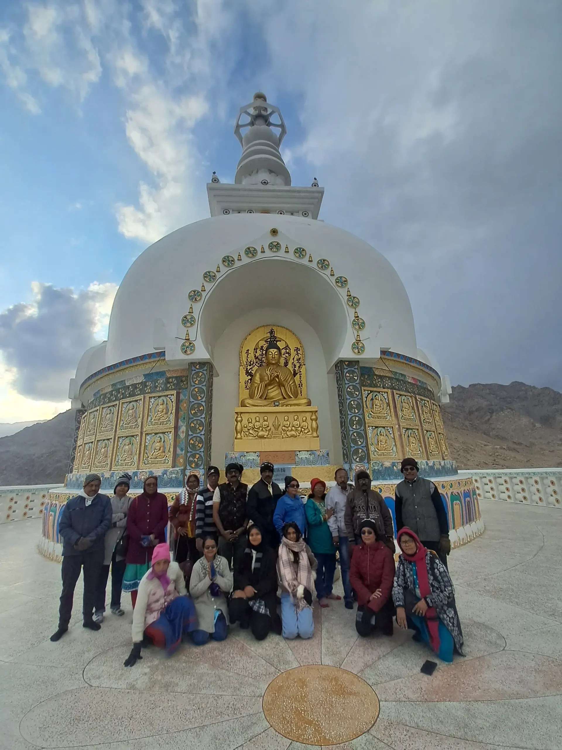 PK Tours travelers in winter jackets posing in front of the white Shanti Stupa and its golden Buddha statue, with brown mountains and a cloudy sky behind, in Leh, Ladakh.